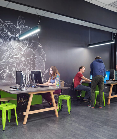 Schüler:innen sitzen auf grünen Plastikhockern an Arbeitsplätzen und Arbeiten am Computer